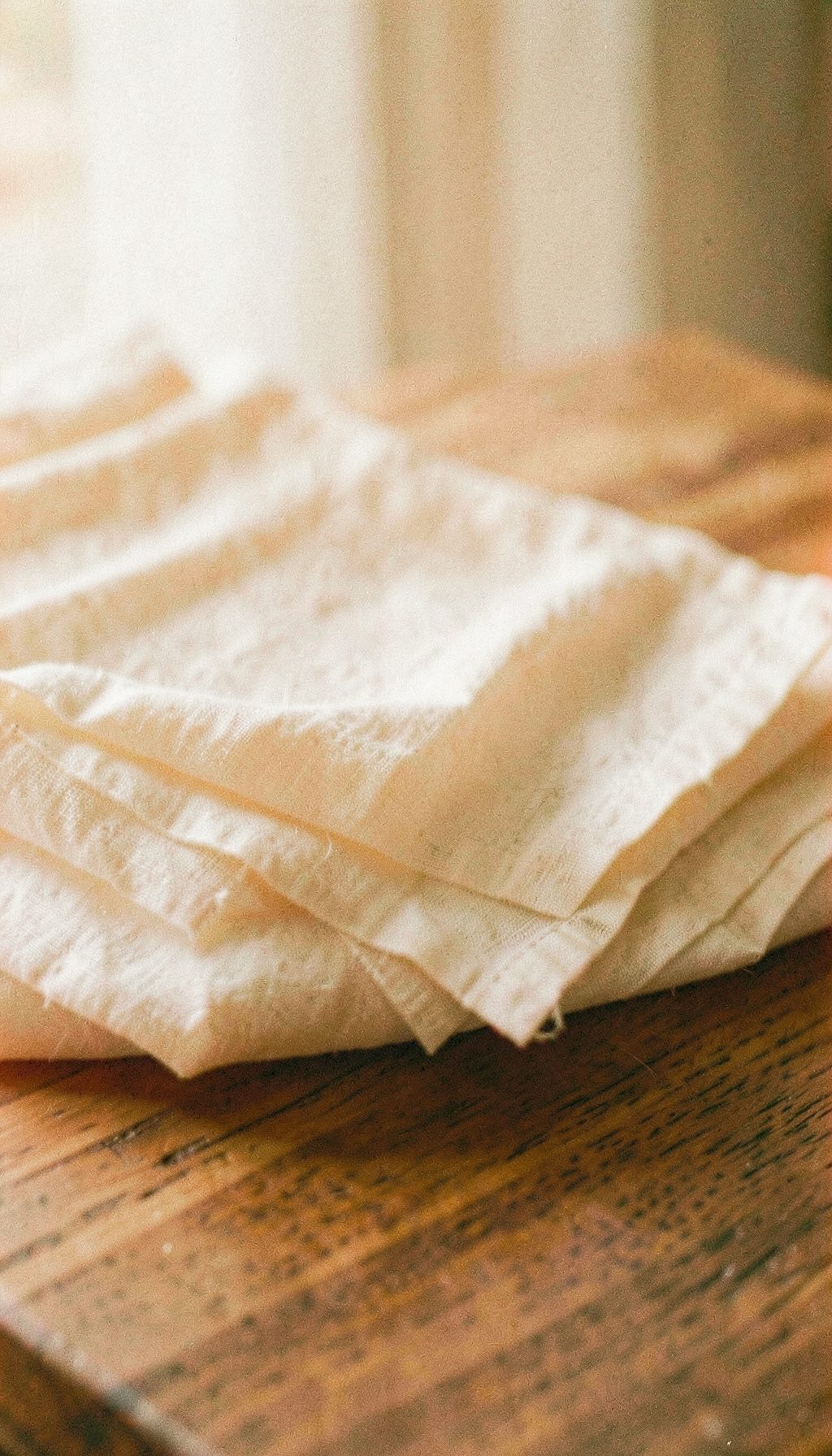 Warm film still of folded linen on a wooden surface in soft daylight, muted cream and beige tones