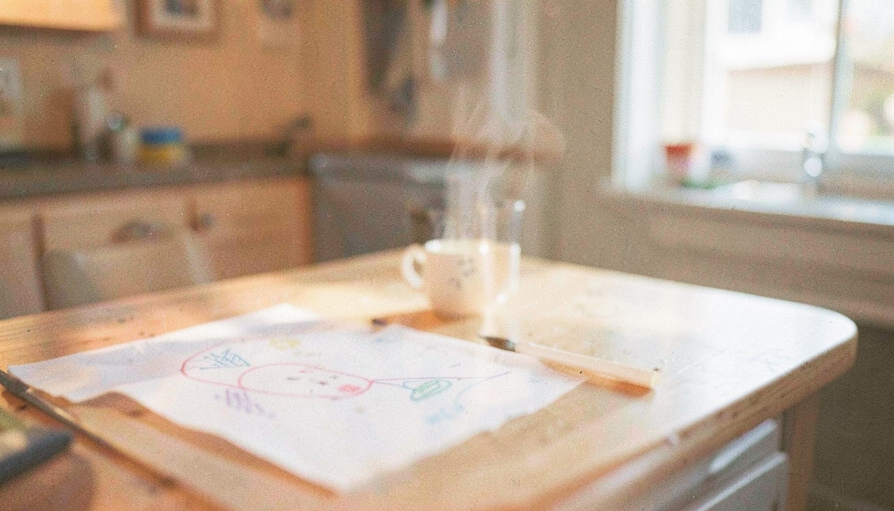 Soft analog film photograph of a sunlit kitchen table with a child's drawing and a steaming mug, cream and pale blue tones, gentle grain