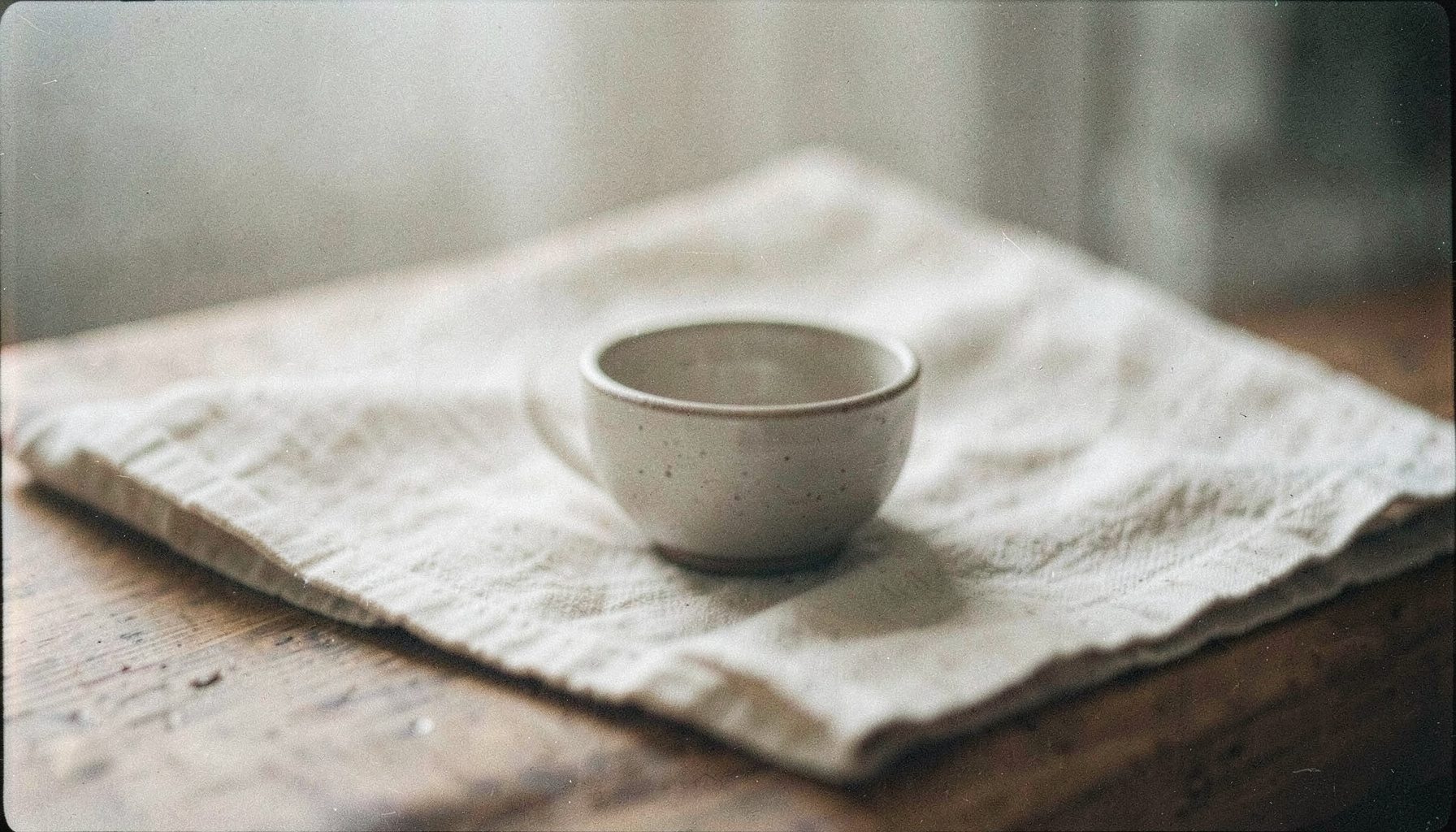 Calm analog film still life with a small ceramic bowl and folded linen on a wooden surface, soft daylight, gentle grain