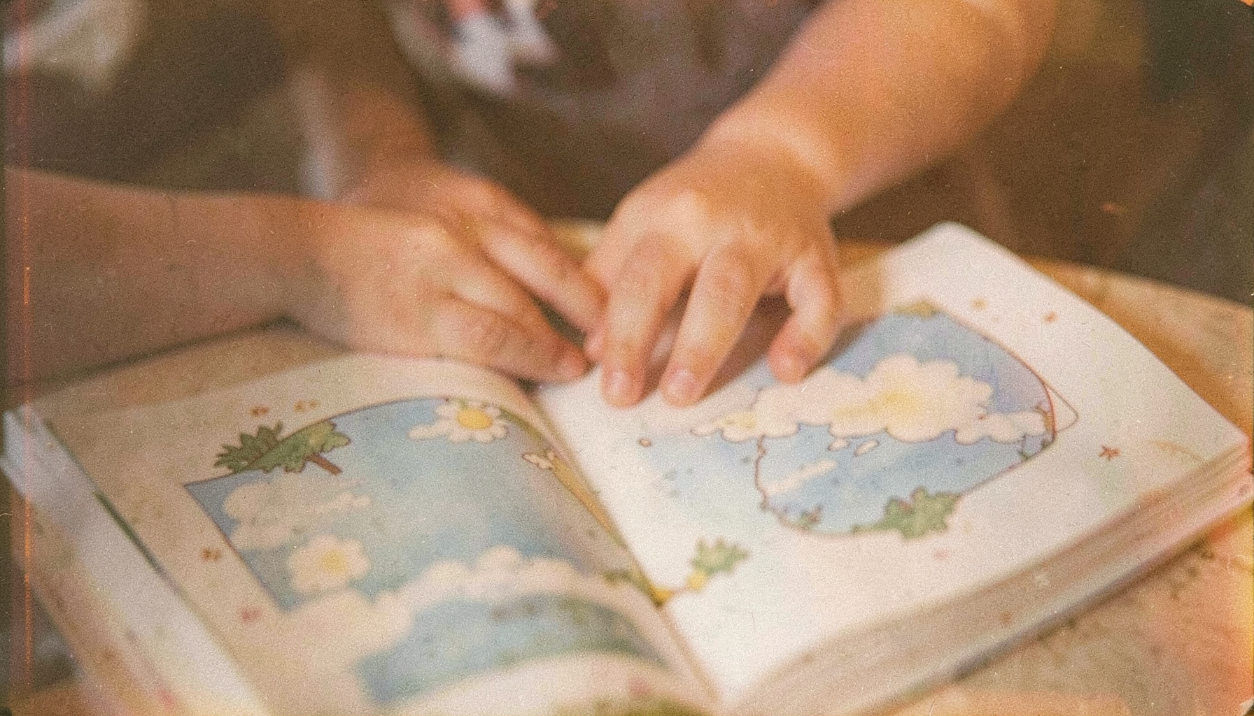 Soft analog film photograph of a child's hands resting on an open picture book about weather, warm muted tones, visible film grain
