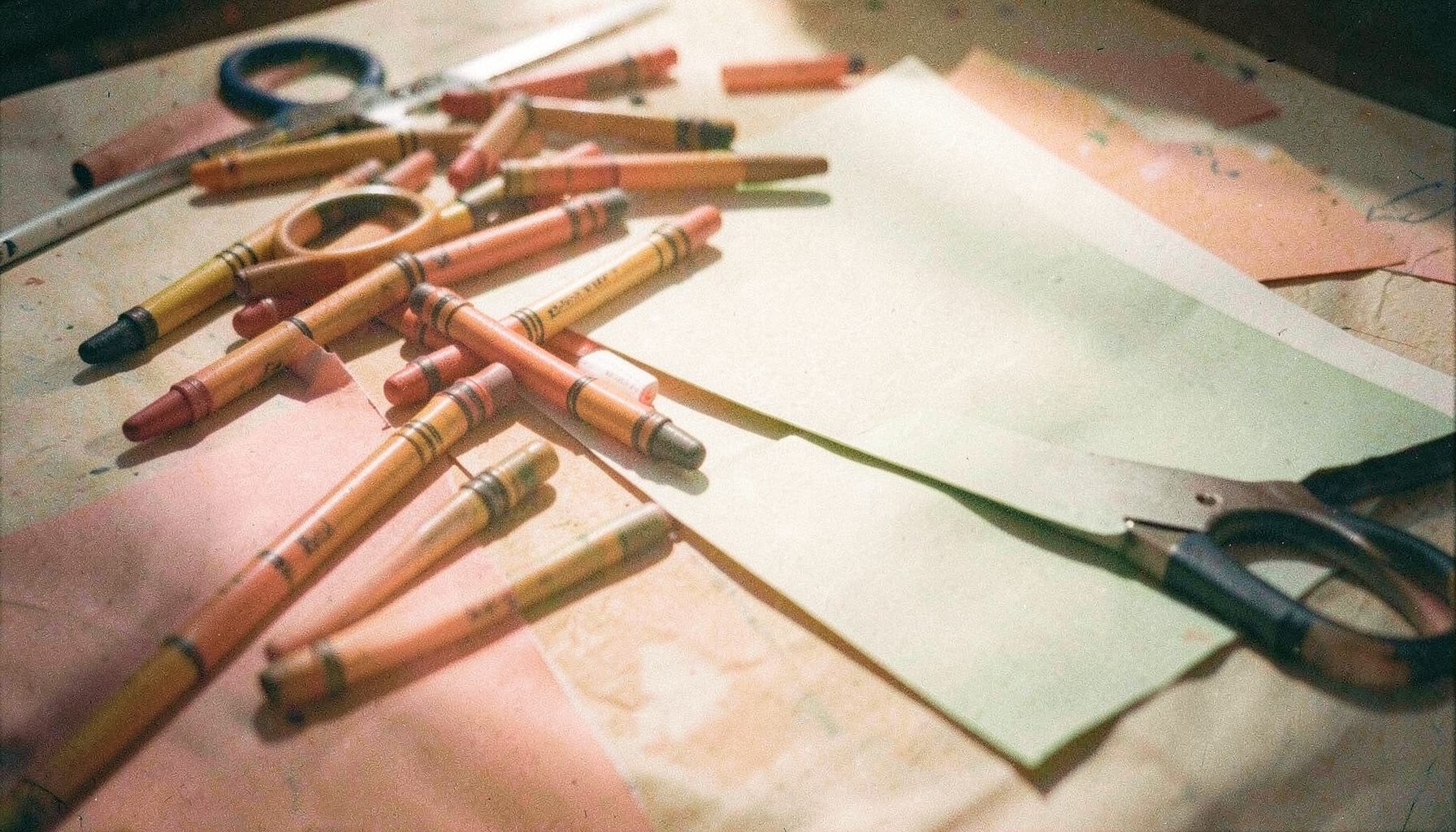Calm analog film still life of crayons, safety scissors, and construction paper spread across a sunlit table, visible film grain and soft shadows.