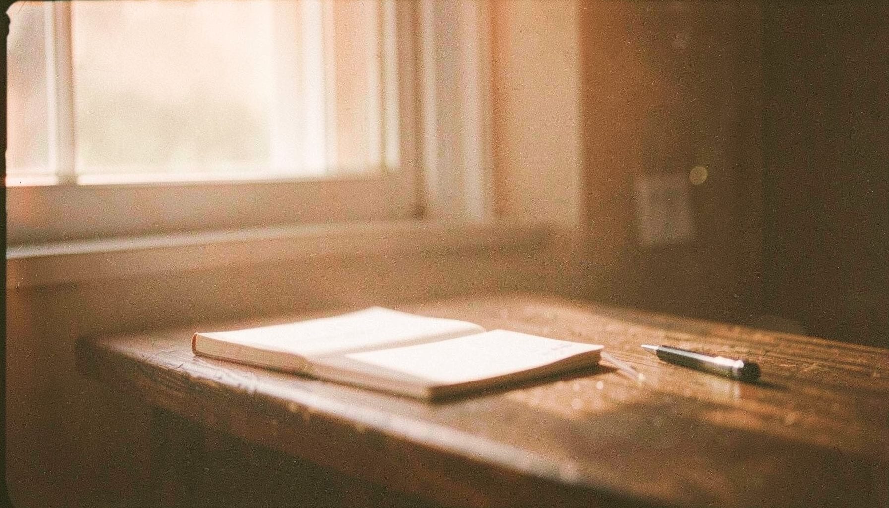 Soft morning light streaming through a window onto a wooden table with a journal and pen, peaceful analog film photograph with visible grain